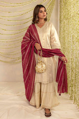 A woman models a traditional Indian outfit consisting of a cream-colored embroidered kurta, matching flared pants, and a deep maroon dupatta with thin gold stripes. She is holding a small gold embroidered handbag and posing in front of a decorated backdrop with gold tinsel drapes.