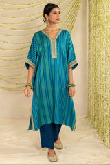 A woman modeling a teal blue and green striped kurta with gold embroidered trim, paired with dark blue pants, standing in front of a decorative gold and cream draped backdrop.