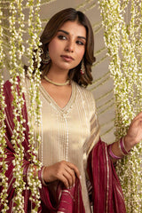 A woman wearing a cream-colored embroidered kurta with a deep V-neckline, paired with a maroon dupatta featuring gold stripes. She is styled with traditional Indian jewelry, including a pearl necklace, earrings, and bangles, and is framed by hanging white flowers.