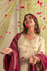 A woman wearing a cream-colored kurta with a deep magenta dupatta with gold stripes, smiling as pink flower petals rain down against a decorated backdrop of hanging gold and white garlands.