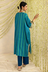 A woman modeling a teal blue and green vertically striped kurta with matching trousers, standing in front of a gold beaded curtain backdrop.