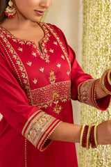A close-up of a woman wearing a red kurta with ornate gold embroidery and matching red-and-gold bangles, posed against a shimmering gold backdrop.