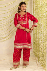A woman modeling a bright red traditional Indian outfit with gold embroidery and patterned pants, standing against a decorated backdrop of draped cream fabric and hanging gold garlands.