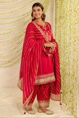 A woman models a traditional Indian outfit consisting of a bright red kurta, matching pants, and a striped red dupatta with gold zari embroidery, set against a decorated backdrop of gold garlands and hanging lights.